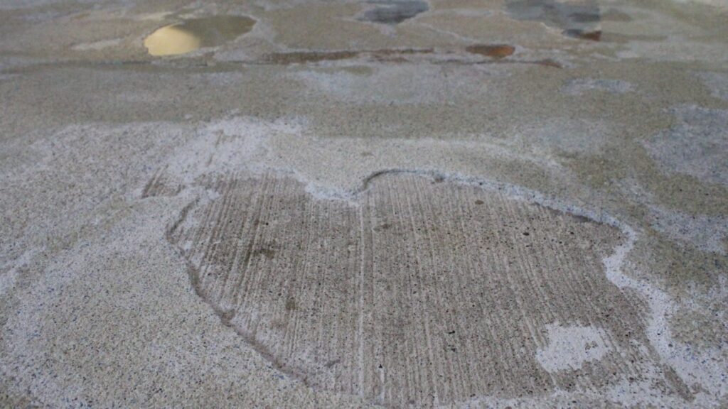Close-up of a textured concrete floor with irregular lighter patches and subtle stains. The surface appears weathered, creating a rustic feel.