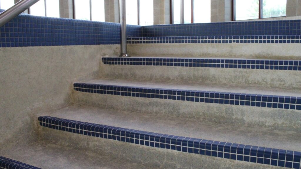 Close-up of wide concrete stairs with blue tiled edges, featuring a silver handrail and large windows in the background.