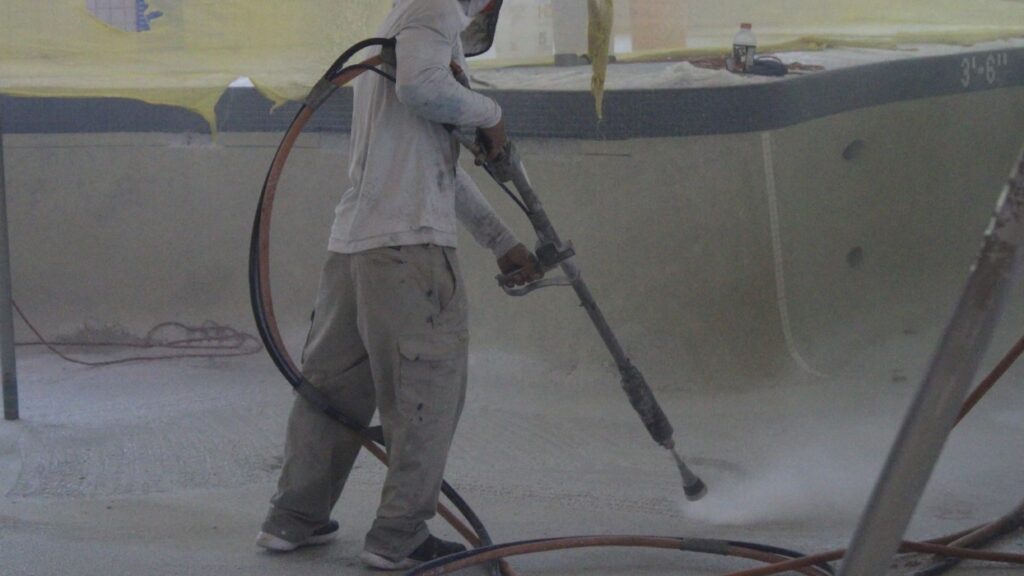 A worker in protective clothing and mask operates a pressure hose, spraying light-colored material on a pool surface. Surrounding area is dusty.