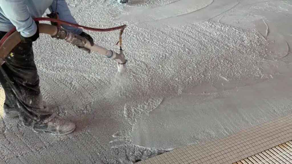Worker applying concrete floor with a large hose, spreading wet mixture evenly. The surface is textured, conveying an ongoing construction process.