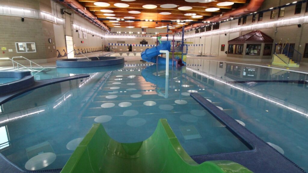 Spacious indoor water park with a large pool featuring a winding green and blue slide, surrounded by modern decor and ceiling lights, creating a playful atmosphere.