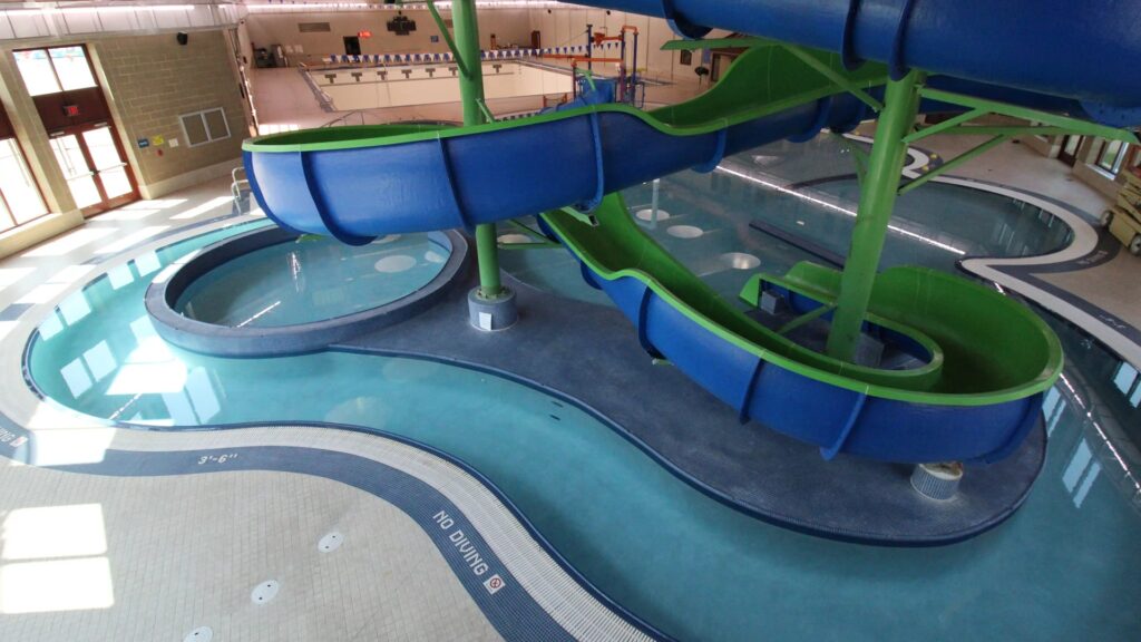 Indoor water park with a blue and green spiral slide leading into a clear pool. Bright lighting and modern design evoke a fun, lively atmosphere.