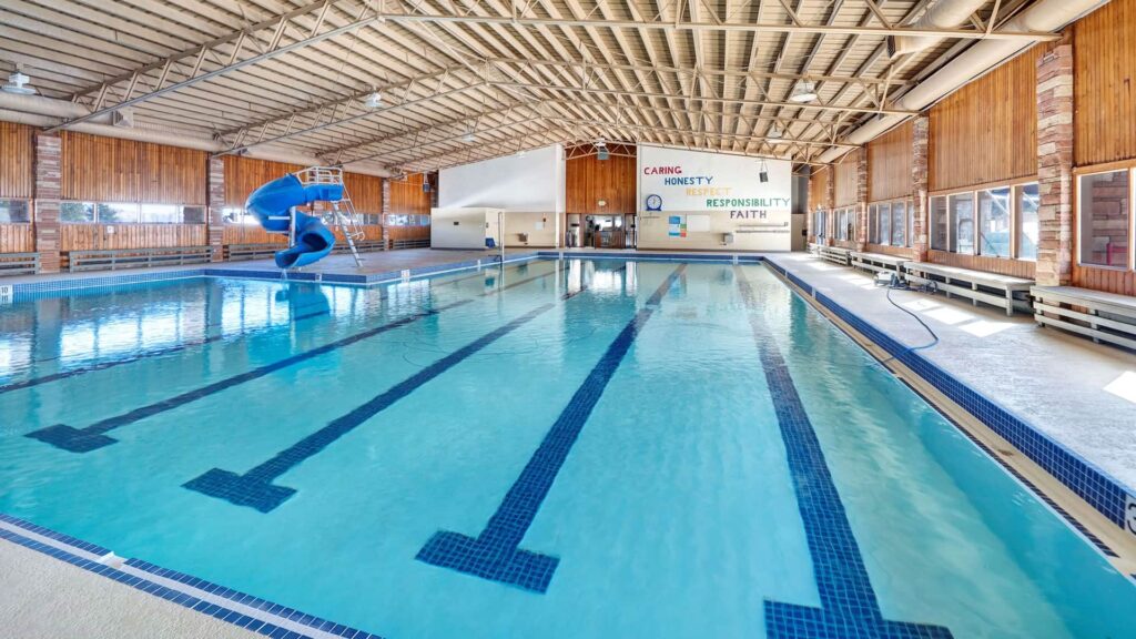 Indoor swimming pool with clear blue water and lane markings. A blue slide is on the left, and a wall displays the words "Caring, Honesty, Responsibility, Faith." Warm lighting and wood paneling create a welcoming atmosphere.