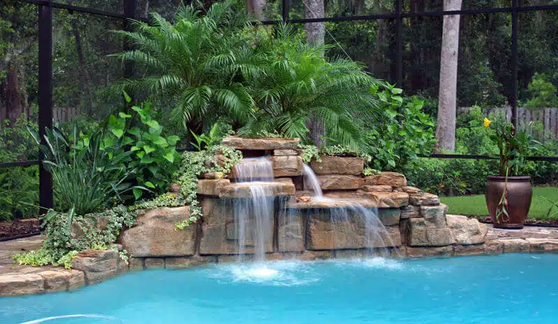Decorative swimming pool waterfall feature with stacked stone design and cascading water surrounded by lush tropical plants — pool landscaping and outdoor renovation inspiration