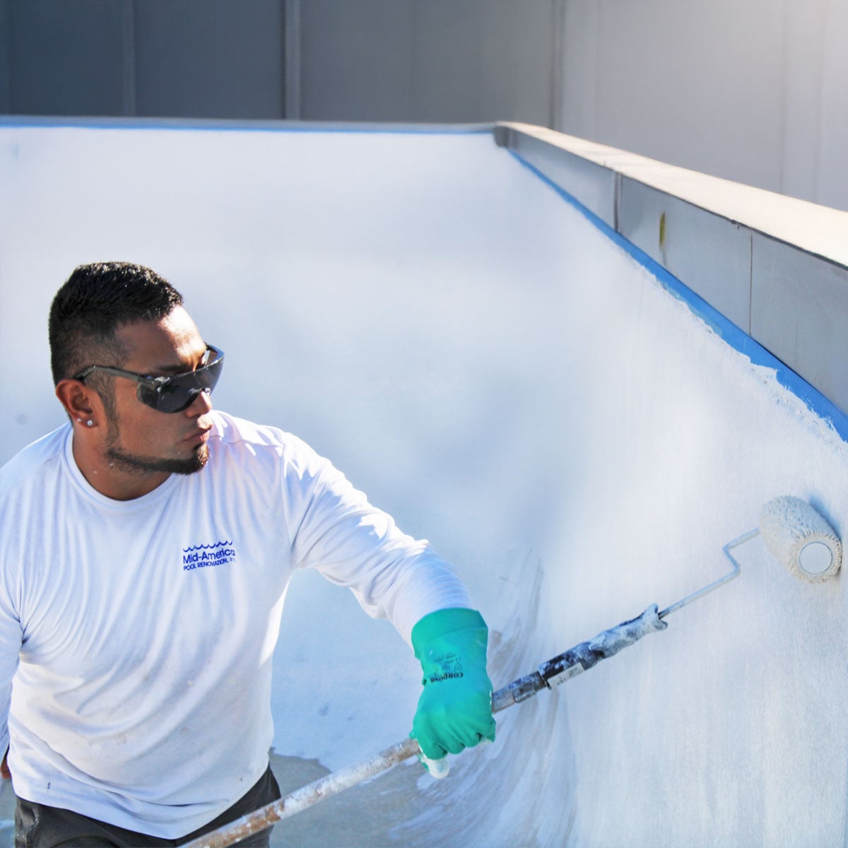 A man wearing sunglasses and green gloves paints the inside of an empty pool with a roller. He appears focused and determined, working under bright sunlight.