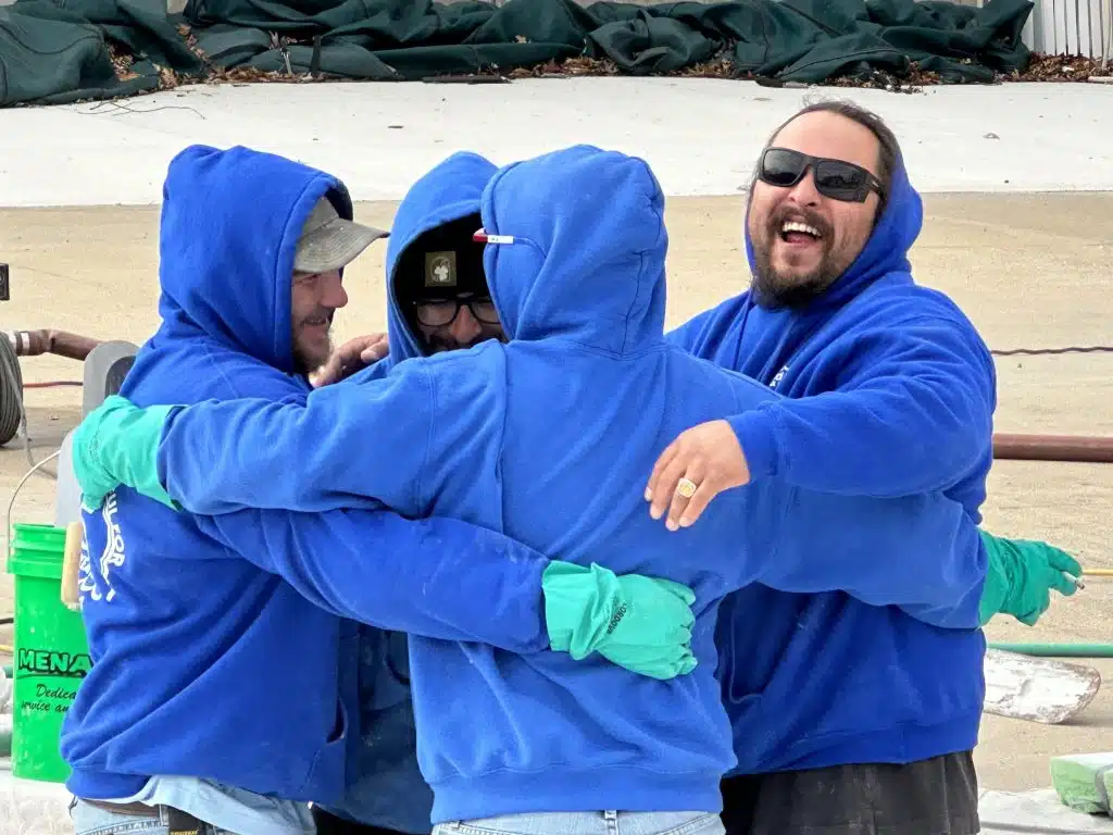 Four people in blue hoodies and green gloves share a joyful group hug outdoors. One person is laughing, adding a sense of camaraderie and happiness.