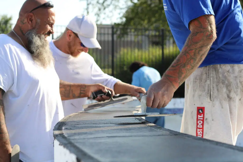 Three men, wearing work clothes, are smoothing concrete with tools on a curved surface. They appear focused and engaged in the outdoor task.