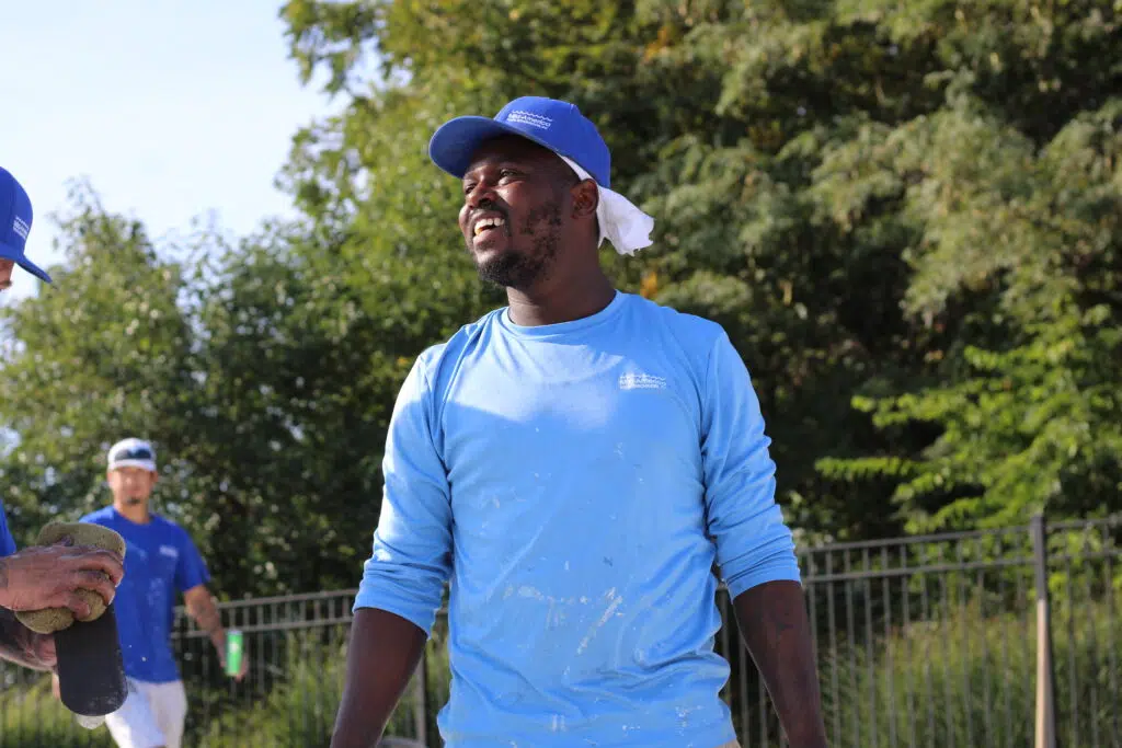 A man in a blue cap and paint-splattered shirt smiles while standing outdoors, with trees and colleagues in similar attire in the background. The mood is cheerful and relaxed.