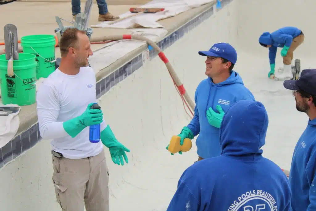 Four workers in blue hoodies and gloves are resurfacing an empty pool. They are laughing and talking, with tools and buckets around them, creating a positive, collaborative atmosphere.