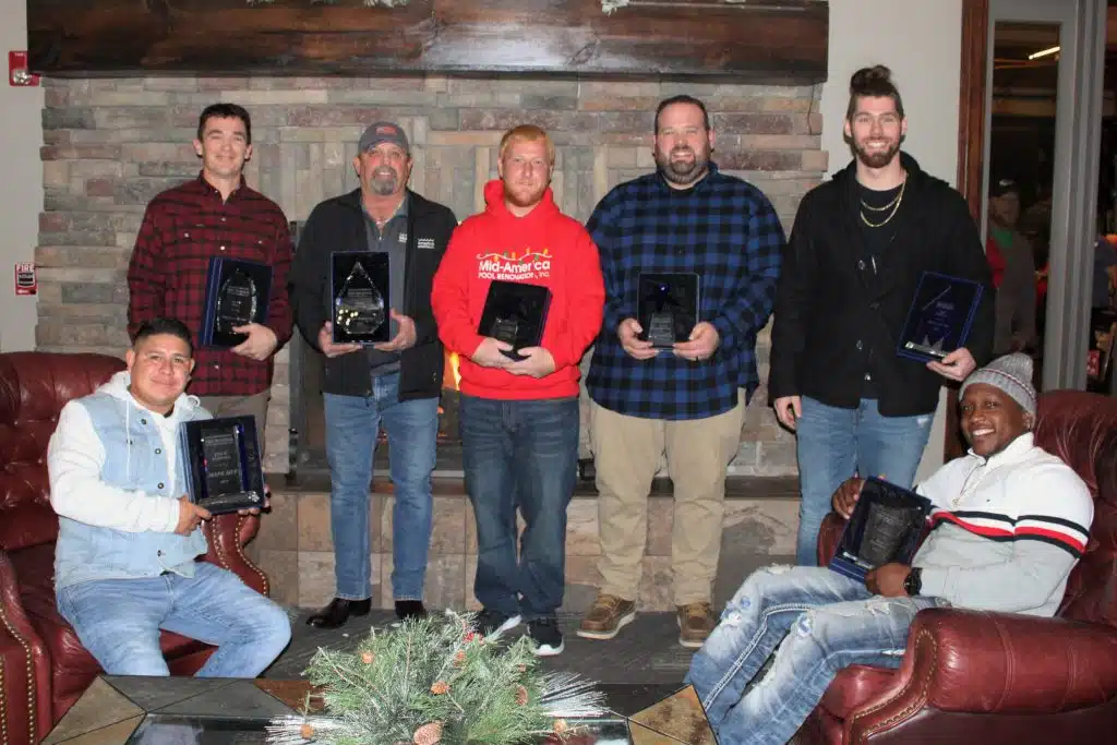 Seven people hold plaques in front of a stone fireplace, suggesting an awards ceremony. They appear relaxed, smiling in casual attire. The room is cozy, conveying a celebratory mood.
