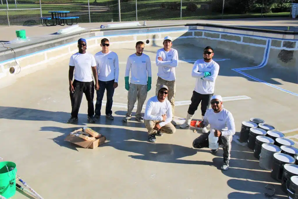 Seven workers in white shirts stand in an empty pool, surrounded by paint buckets. The mood is industrious and collaborative under sunny skies.