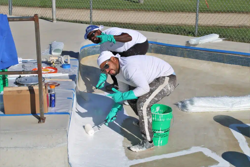 Two people in white attire and gloves paint a skate park bowl. They are focused and smiling, surrounded by paint tools and materials under sunny skies.