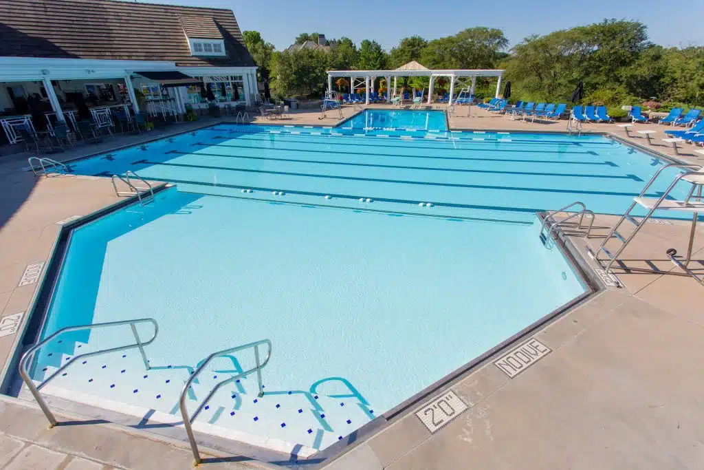 Outdoor pool with lap lanes and shallow steps, surrounded by lounge chairs and trees. Adjacent buildings suggest a relaxed, resort-like setting.
