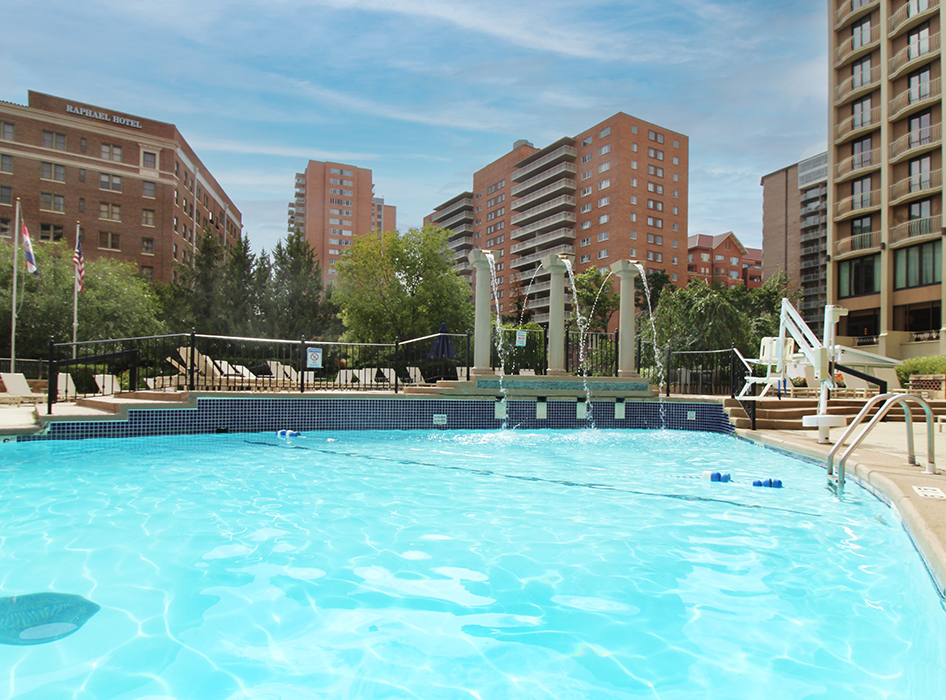Bright outdoor swimming pool with clear blue water, lounge chairs, and red‑brick apartment buildings in background — residential or hotel pool renovation and recreational facility design