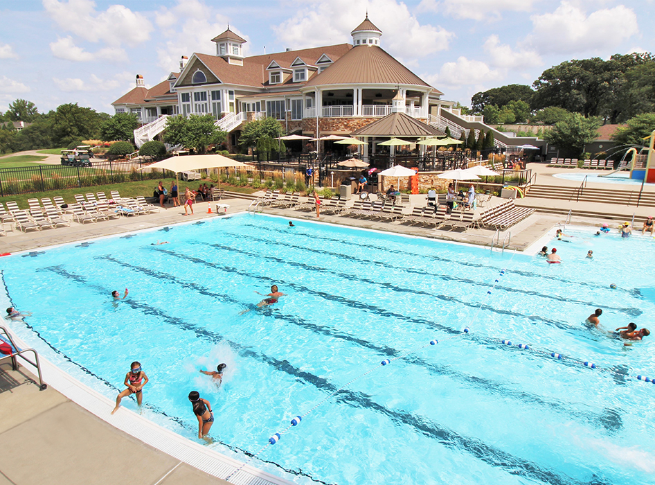 Outdoor pool area with swimmers enjoying a sunny day. A large clubhouse with windows and turrets is visible in the background, surrounded by lounge chairs.