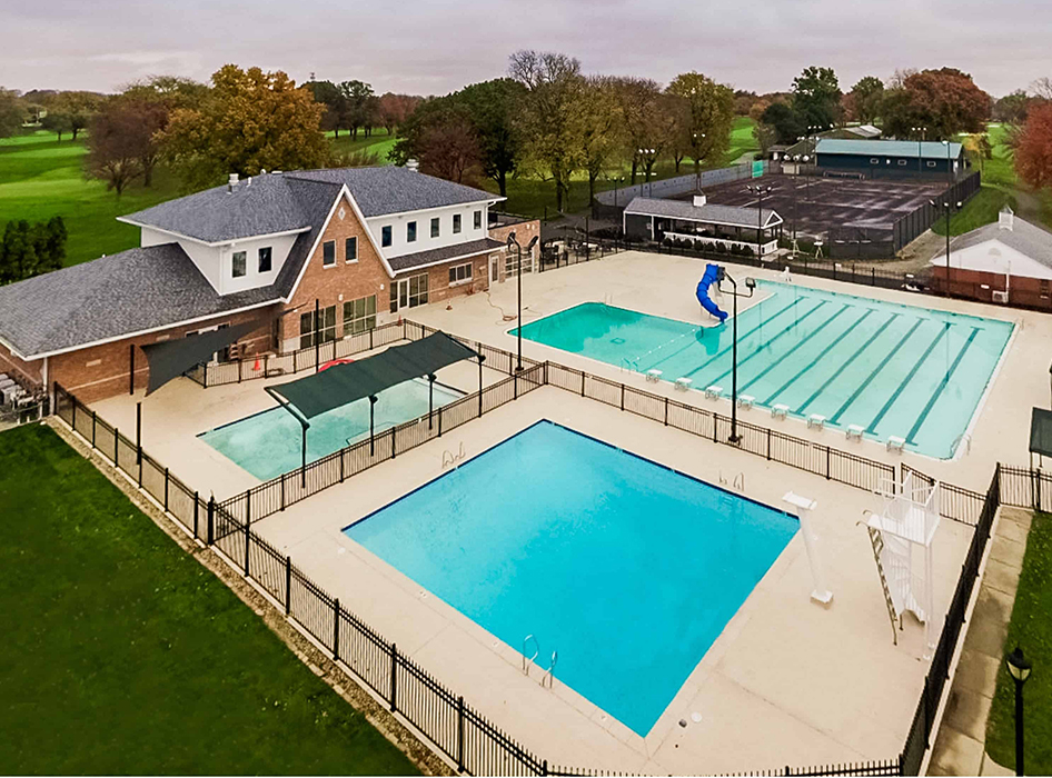 Outdoor recreational area with two swimming pools, a brick clubhouse, and tennis courts. Trees surround the area, conveying a peaceful vibe.