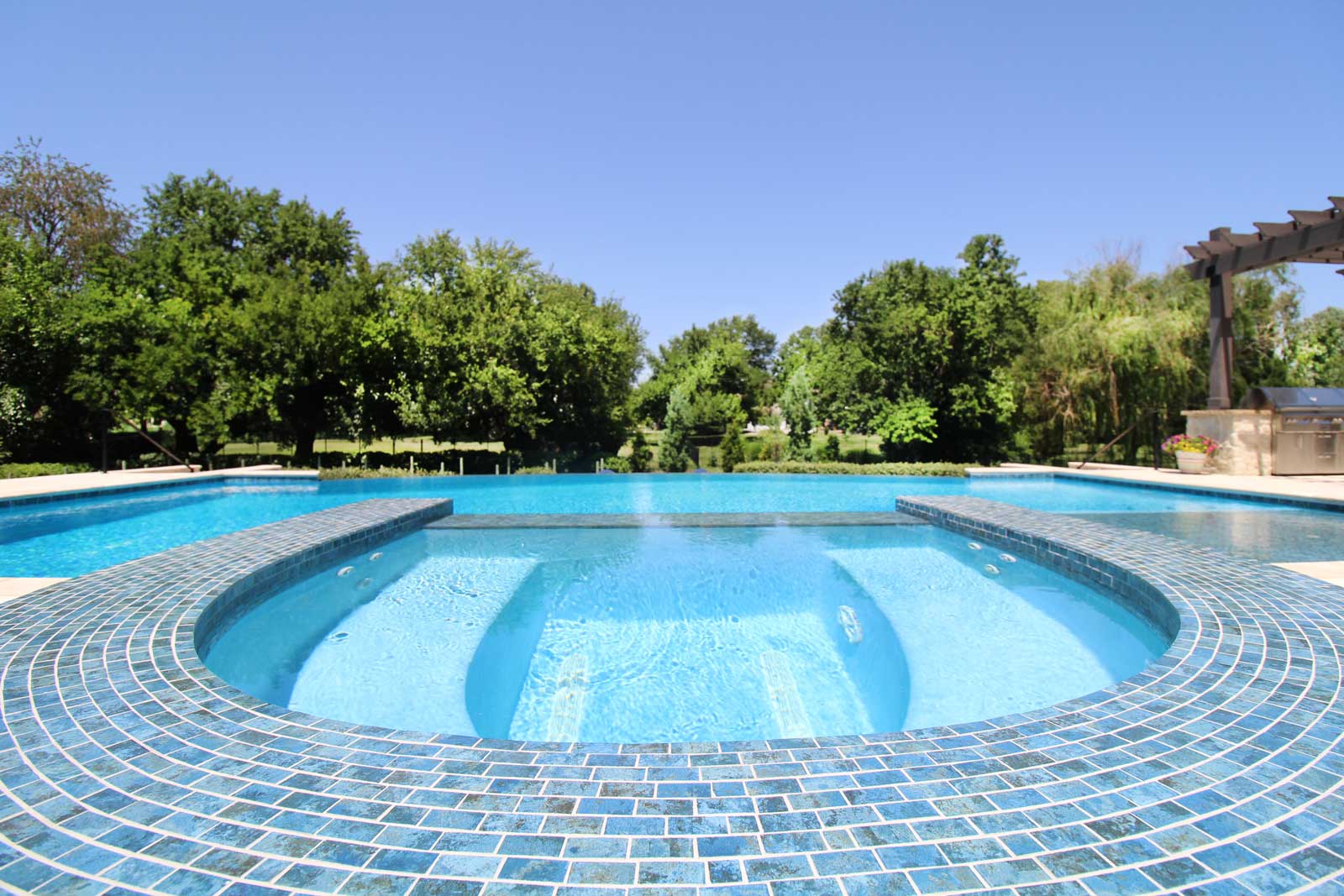 Luxurious swimming pool with a circular hot tub, surrounded by lush green trees under a clear blue sky, conveying a serene and inviting atmosphere.