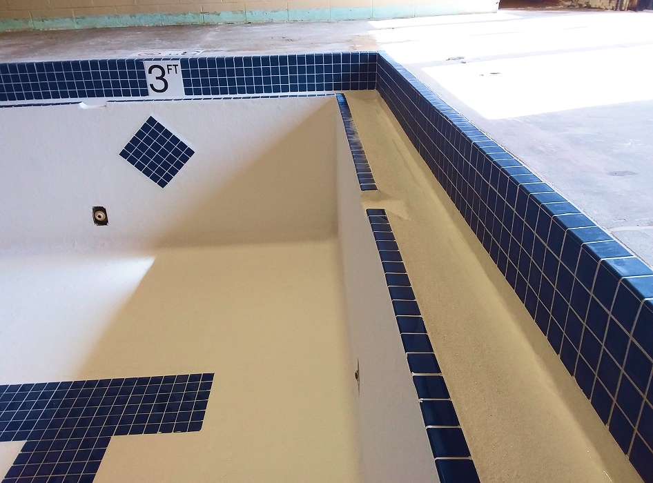 Empty swimming pool with blue-tiled edges and floor. A sign reads "3 FT." Sunlight casts shadows, giving a calm, abandoned feel.