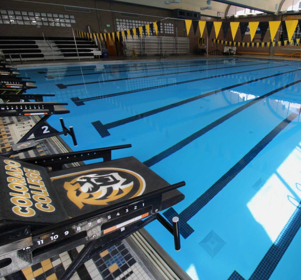 Indoor swimming pool with five marked lanes, surrounded by yellow and black flags. Diving platforms display college branding. Sunlight reflects on the water.