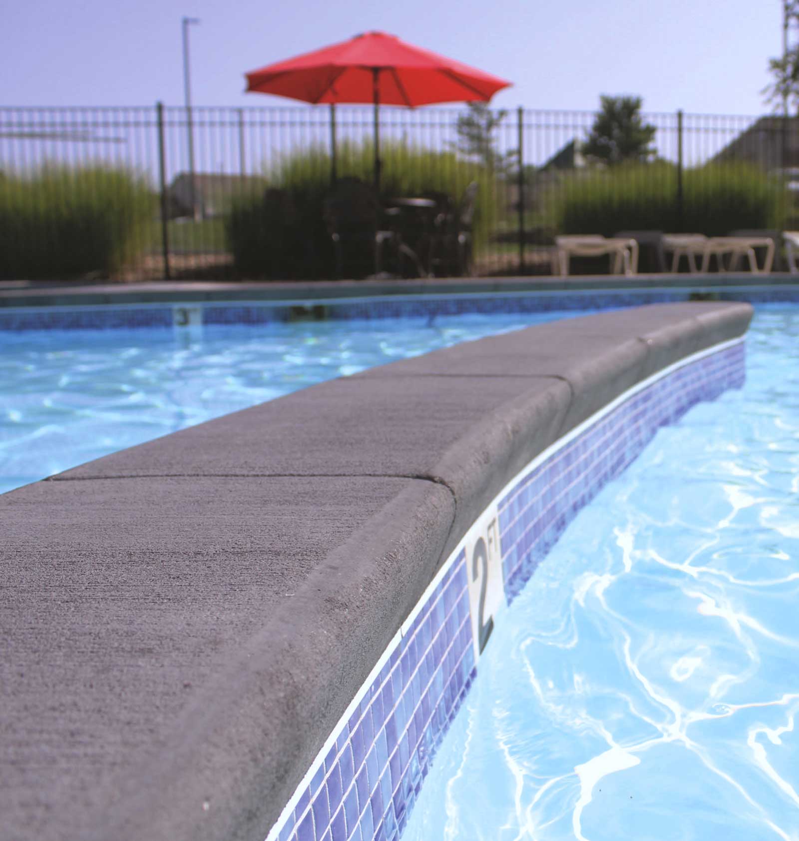 Close-up of a swimming pool edge with blue mosaic tiles and water level marked at 2 feet. A red umbrella shades a table, creating a relaxed vibe.