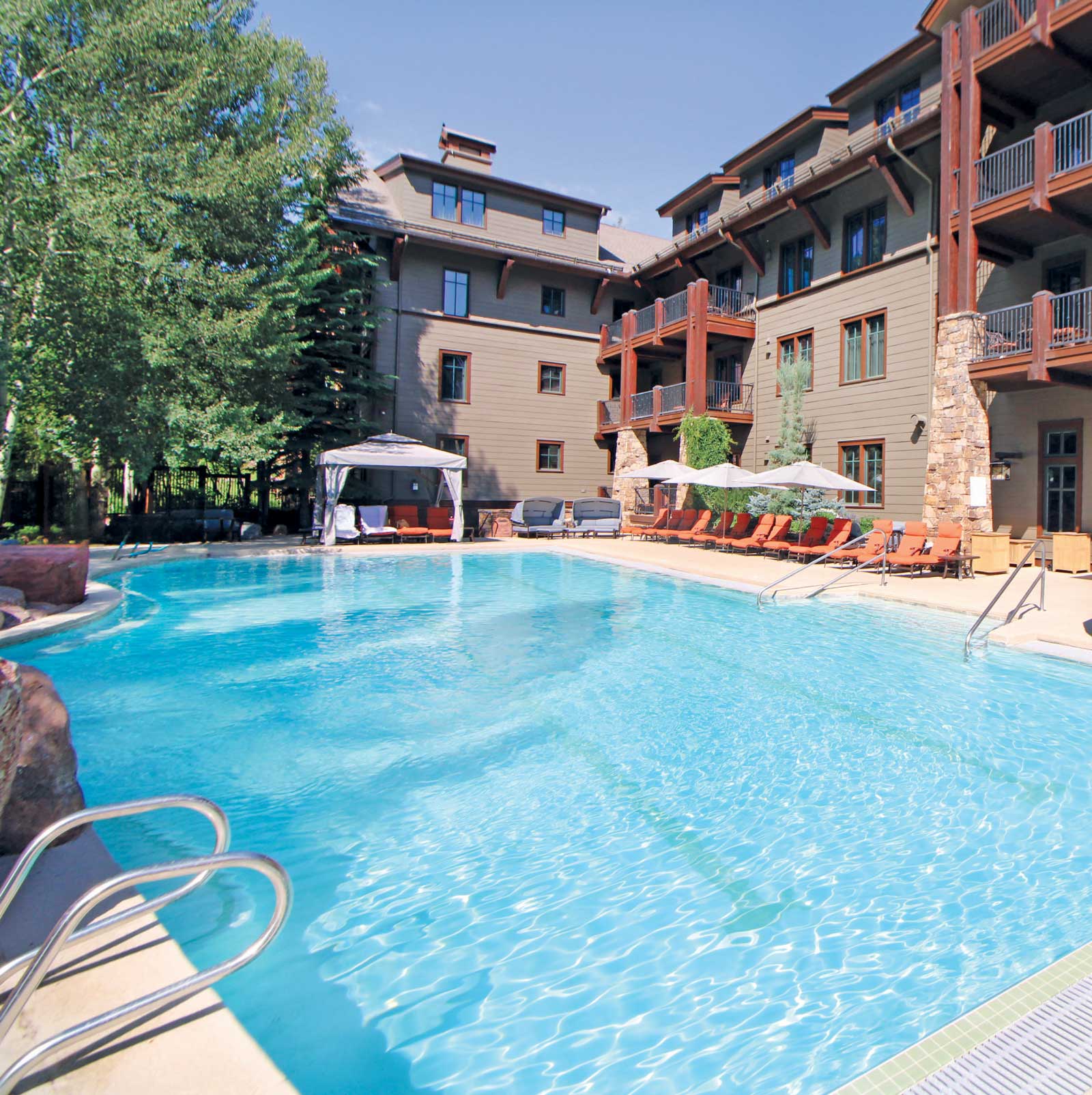 Outdoor pool area at a rustic lodge with wooden balconies, red lounge chairs, and umbrellas. A cabana is shaded by trees beside the building. Calm and inviting.