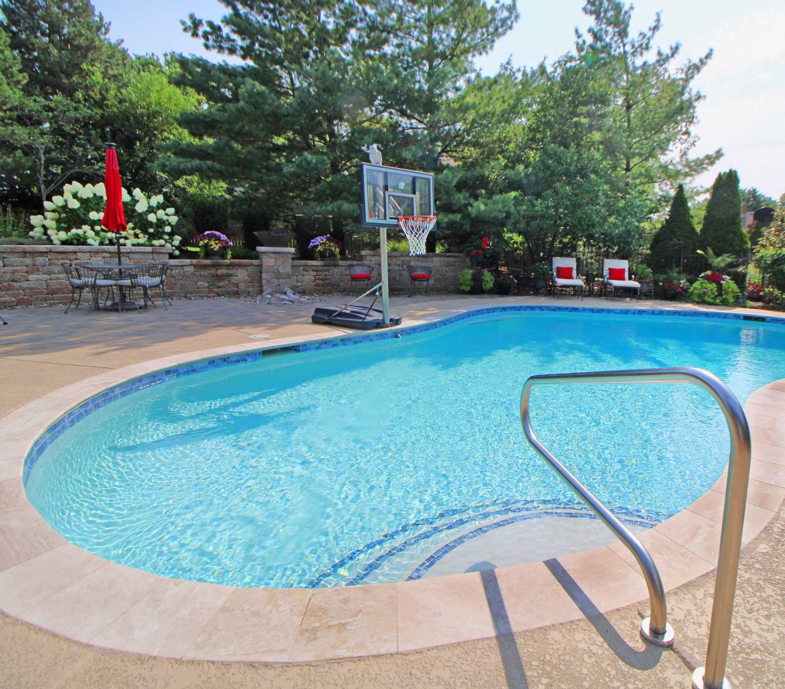 A serene backyard pool with clear blue water, flanked by a basketball hoop. Surrounded by lush greenery, red chairs, and a patio set with an umbrella.