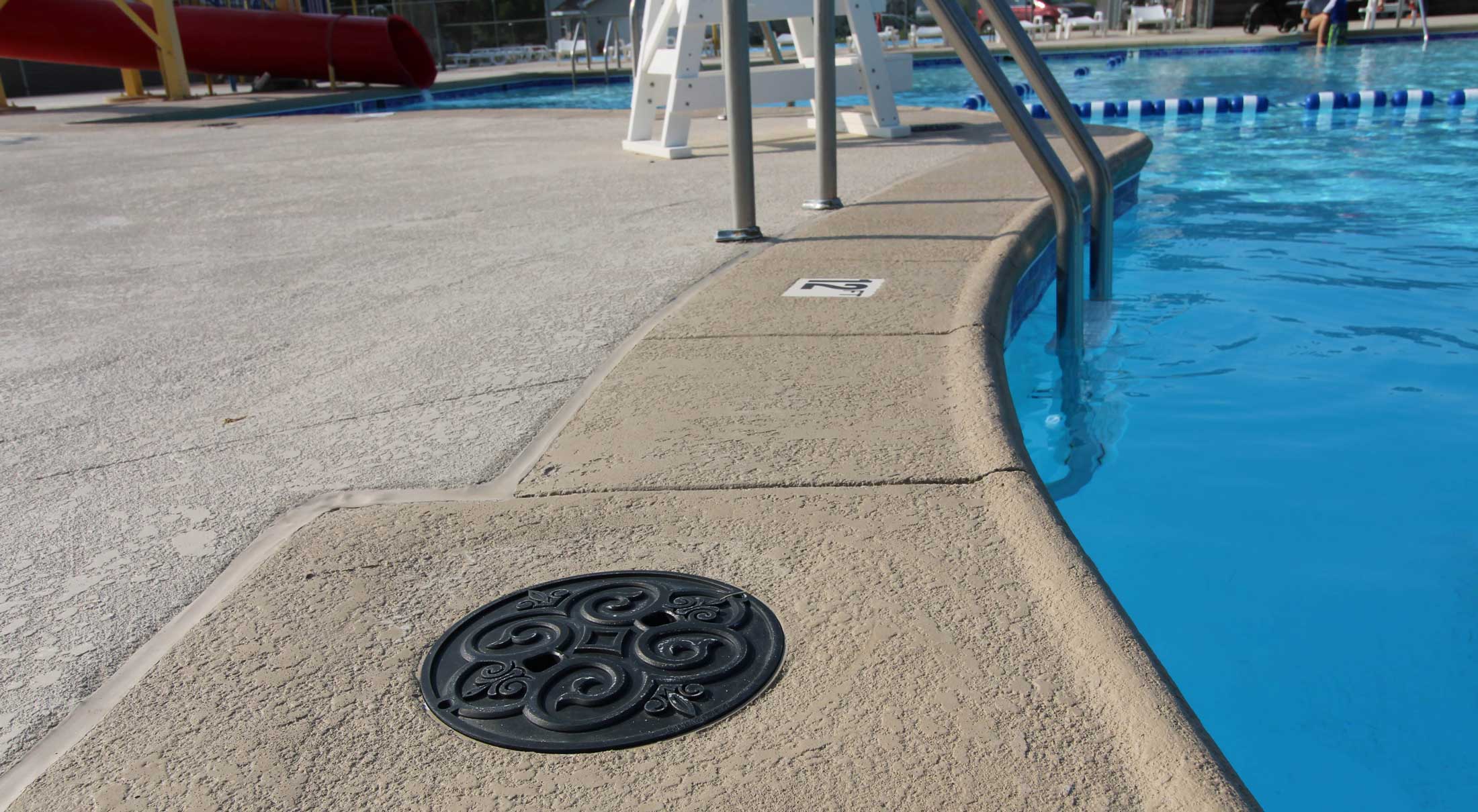View of a poolside with clear blue water. A decorative drain cover and a pool ladder are visible on the beige deck. A red slide is in the background.