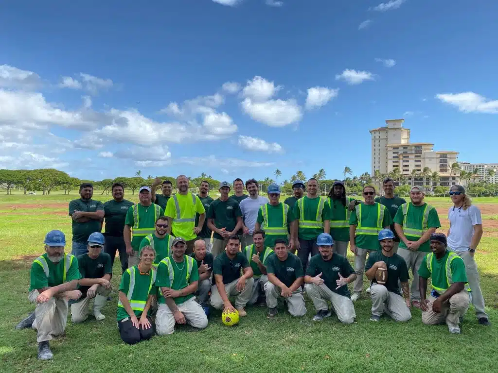 A diverse group of 30 people wearing green shirts and safety vests poses outdoors on a grassy field under a blue sky. A sense of camaraderie is felt.