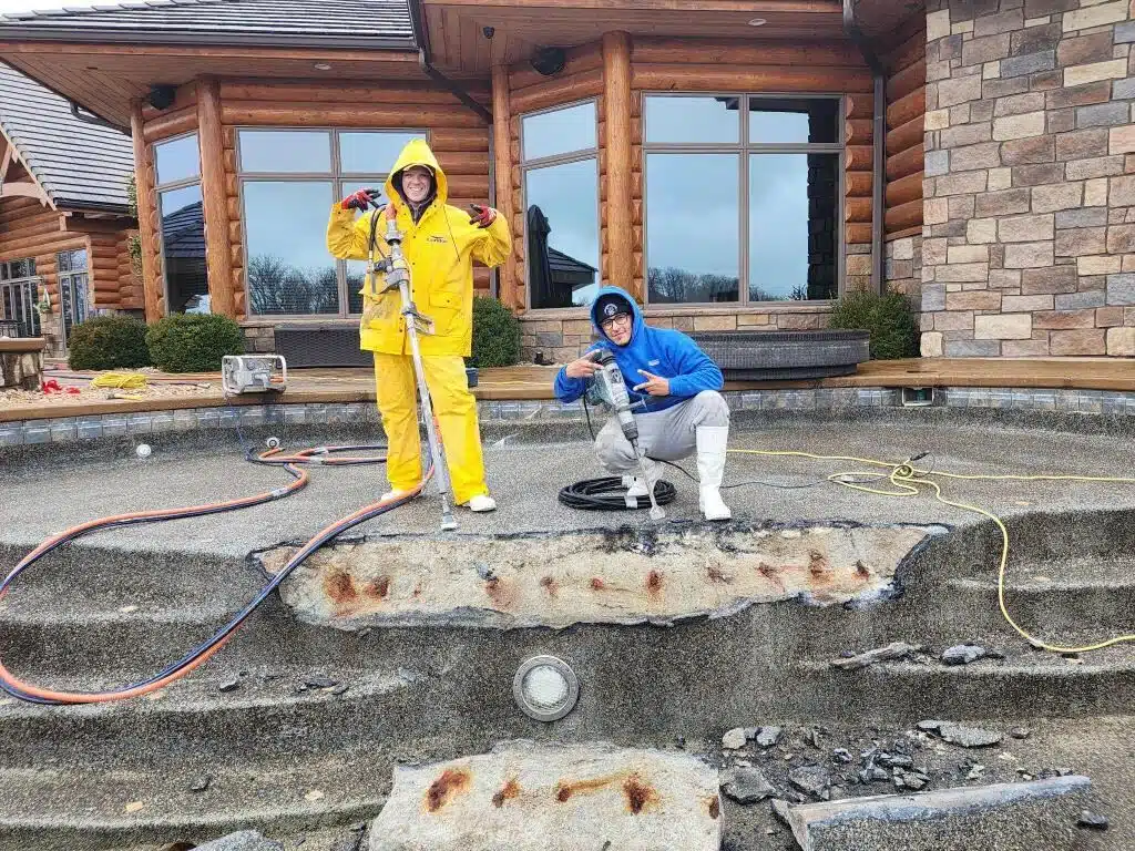Two people pose on an empty, damaged pool. One in a yellow rain suit stands with tools; the other kneels in a blue hoodie. The mood is playful.