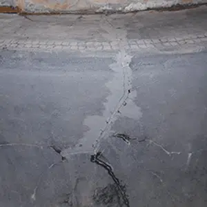 A cracked gray concrete surface with jagged lines running across it. The top edge has a rough, textured finish, suggesting wear and deterioration.