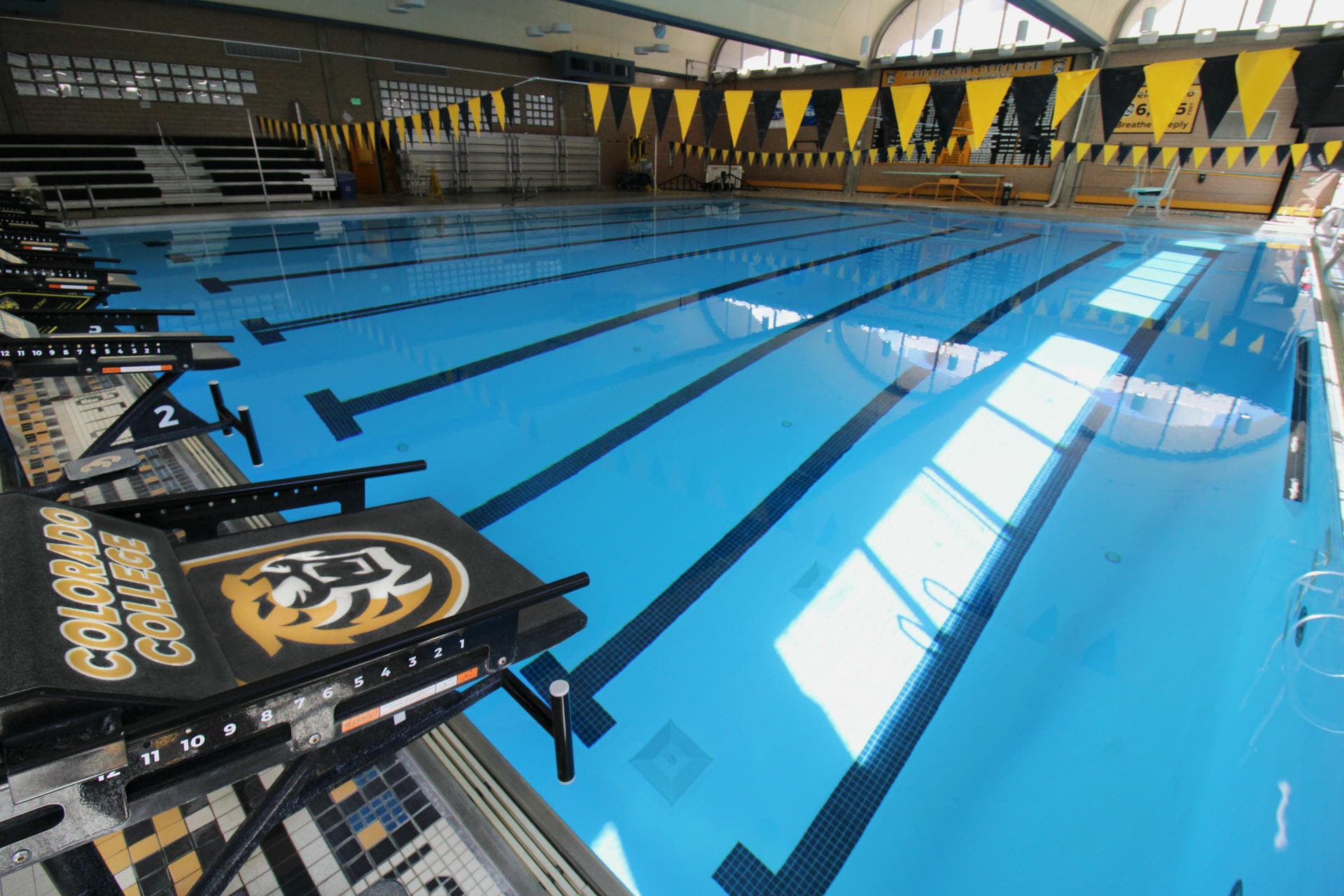 Indoor swimming pool with five marked lanes, surrounded by yellow and black flags. Diving platforms display college branding. Sunlight reflects on the water.