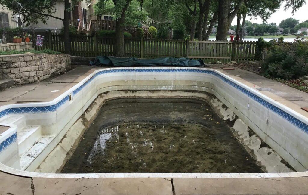 An empty, neglected swimming pool with dirty water at the bottom, surrounded by overgrown foliage and a wooden fence. The mood is abandoned and unkempt.