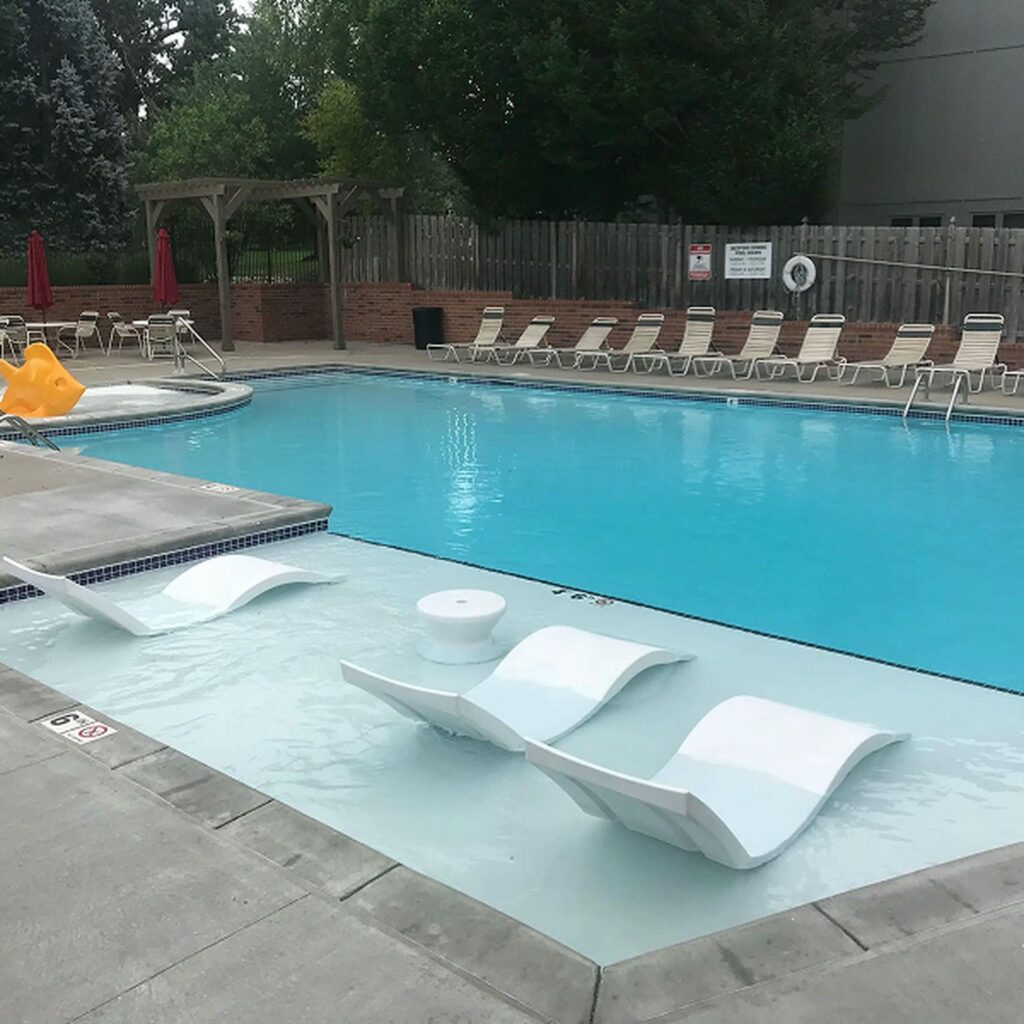 Outdoor pool area with light blue water, surrounded by lounge chairs and red umbrellas. White in-water loungers are in the shallow end. Serene and inviting atmosphere.