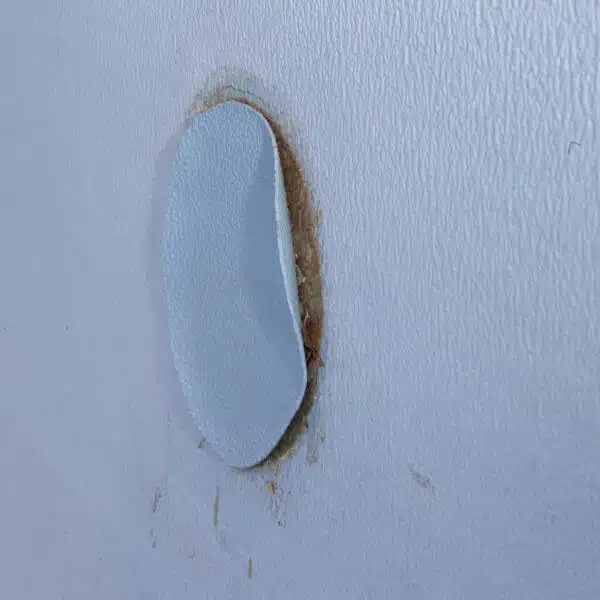 A beige wall with a rough, oval-shaped patch slightly peeling off, revealing a brownish area underneath. The image conveys a sense of disrepair.