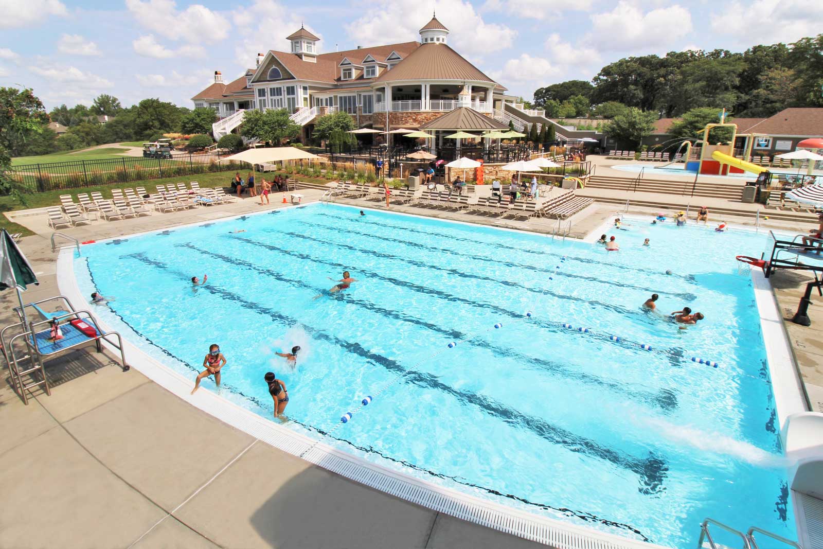 A large outdoor swimming pool with lanes and people enjoying the water. Surrounding the pool are lounge chairs and a clubhouse in the background under a blue sky.
