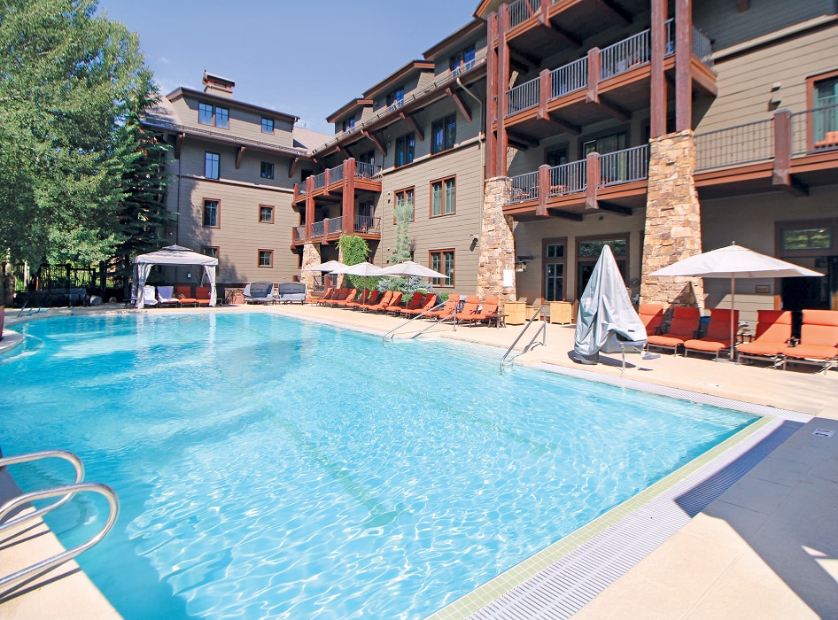 Outdoor pool at a resort with clear blue water. Surrounded by red lounge chairs, umbrellas, and a rustic building in the background. Sunny day.