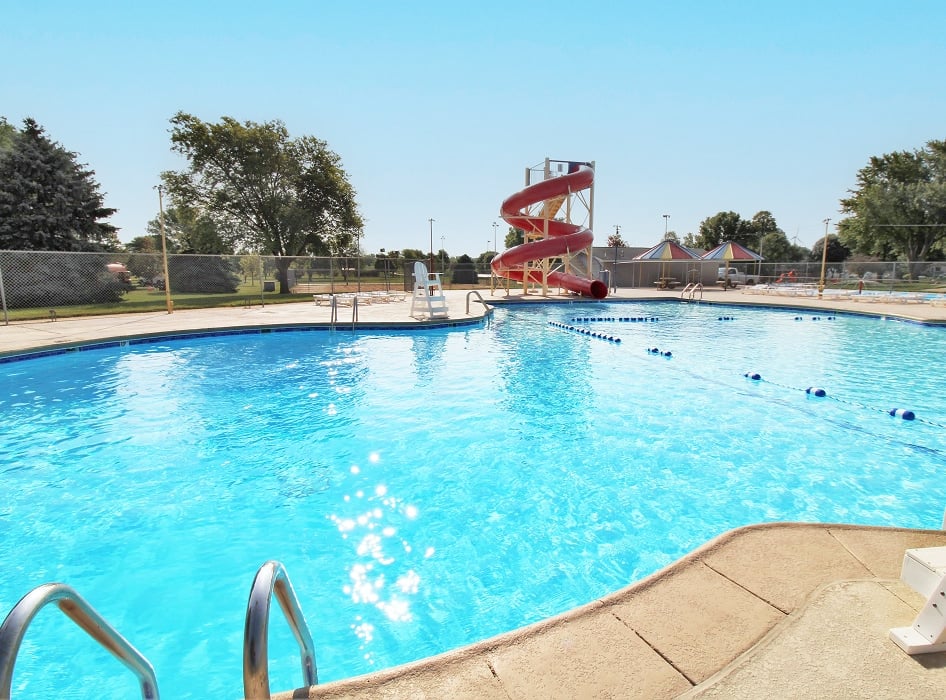 Backyard with a rectangular pool, waterslide, and bubbling fountain. Surrounded by a patio, garden, and trees under a clear blue sky. Tranquil setting.