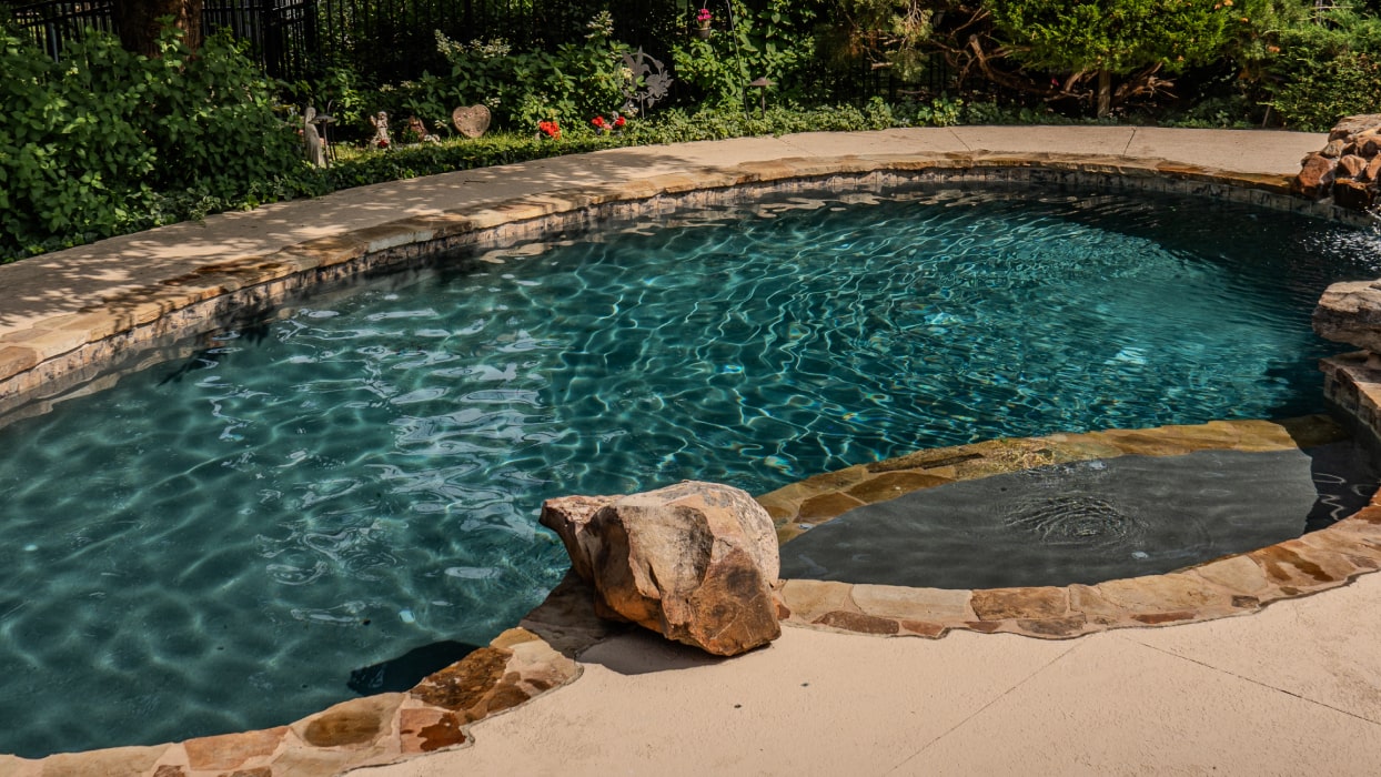 A small, oval-shaped swimming pool with clear, rippling water is surrounded by a stone patio. Lush greenery and a few colorful flowers are in the background, providing a serene and inviting atmosphere.