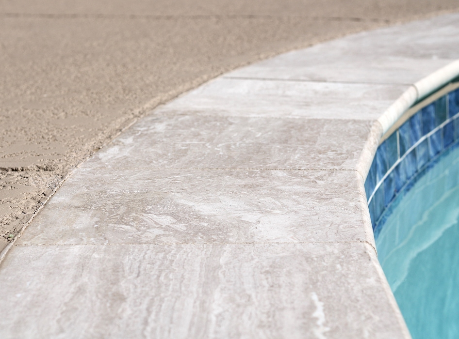 Curved pool edge with light gray tiles leading into aqua blue water. Surrounding area is sandy with a smooth finish, conveying a serene atmosphere.