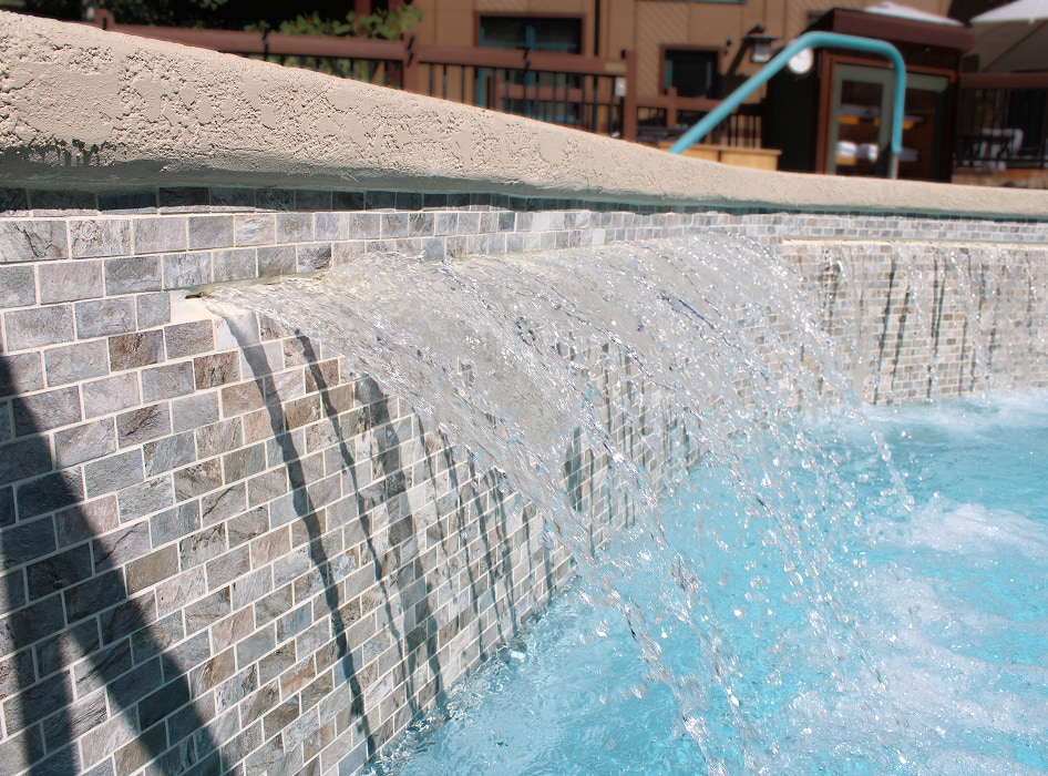 Water cascades over a tiled ledge into a pool, creating a relaxing and refreshing scene. The clear blue water contrasts with the mosaic tiles.