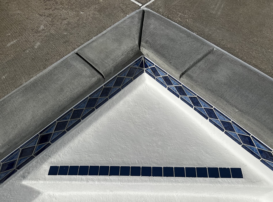 Close-up of a pool corner with gray concrete coping. Blue and white mosaic tiles form a border. The clean design evokes simplicity and precision.