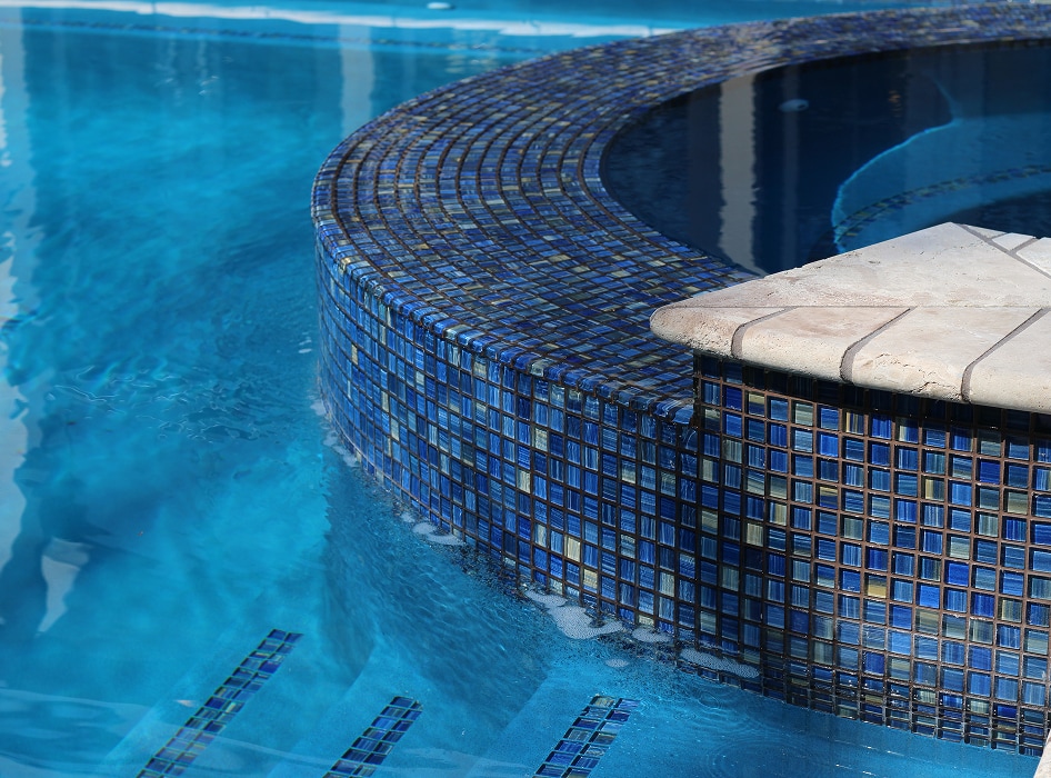 A close-up of a round, tiled jacuzzi with deep blue mosaic tiles. It merges seamlessly into a clear blue pool, creating a serene and luxurious atmosphere.