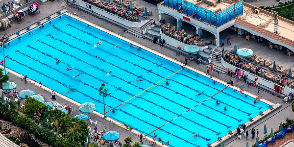 A large outdoor swimming pool with swimmers in lanes, surrounded by lounging areas with umbrellas and chairs. The scene is lively and bustling.