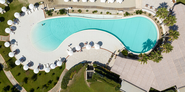 Aerial view of a large, uniquely shaped swimming pool surrounded by white umbrellas and lounge chairs. Green grass and palm trees enhance the resort ambiance.