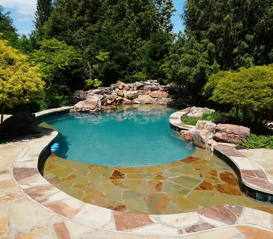 A serene backyard pool with clear blue water surrounded by natural stone tiles. Lush greenery and a rock waterfall add a tranquil, inviting atmosphere.