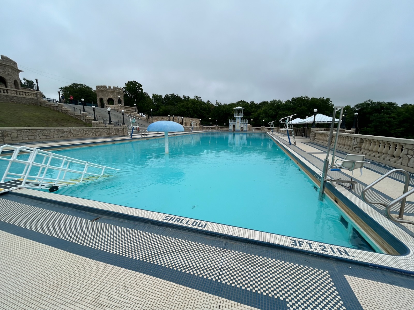 A large, empty swimming pool with clear blue water, surrounded by a stone deck and greenery in the background. The sky is overcast.