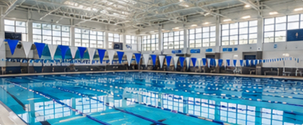 Spacious indoor swimming pool with clear blue water, lined with lane dividers. Bright lighting and large windows create an inviting and airy atmosphere.