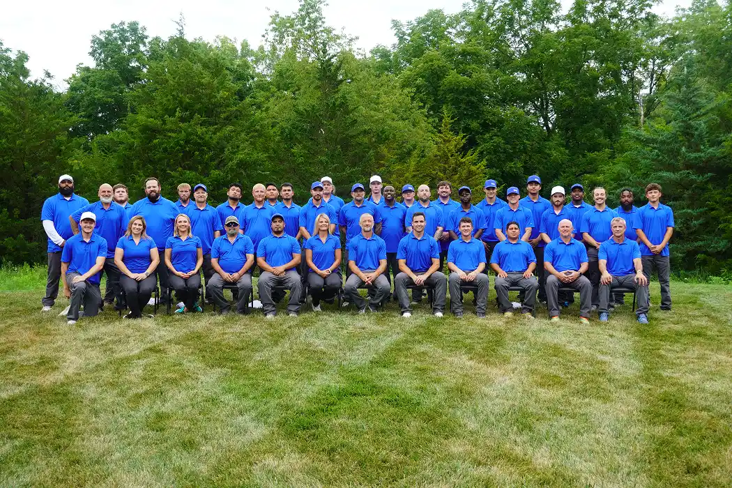 A large group of people, all wearing blue shirts and gray pants, standing and sitting together outdoors in a grassy area surrounded by trees.