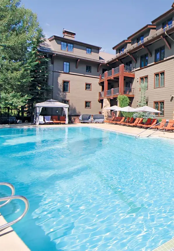 Outdoor pool area at a rustic lodge with wooden balconies, red lounge chairs, and umbrellas. A cabana is shaded by trees beside the building. Calm and inviting.