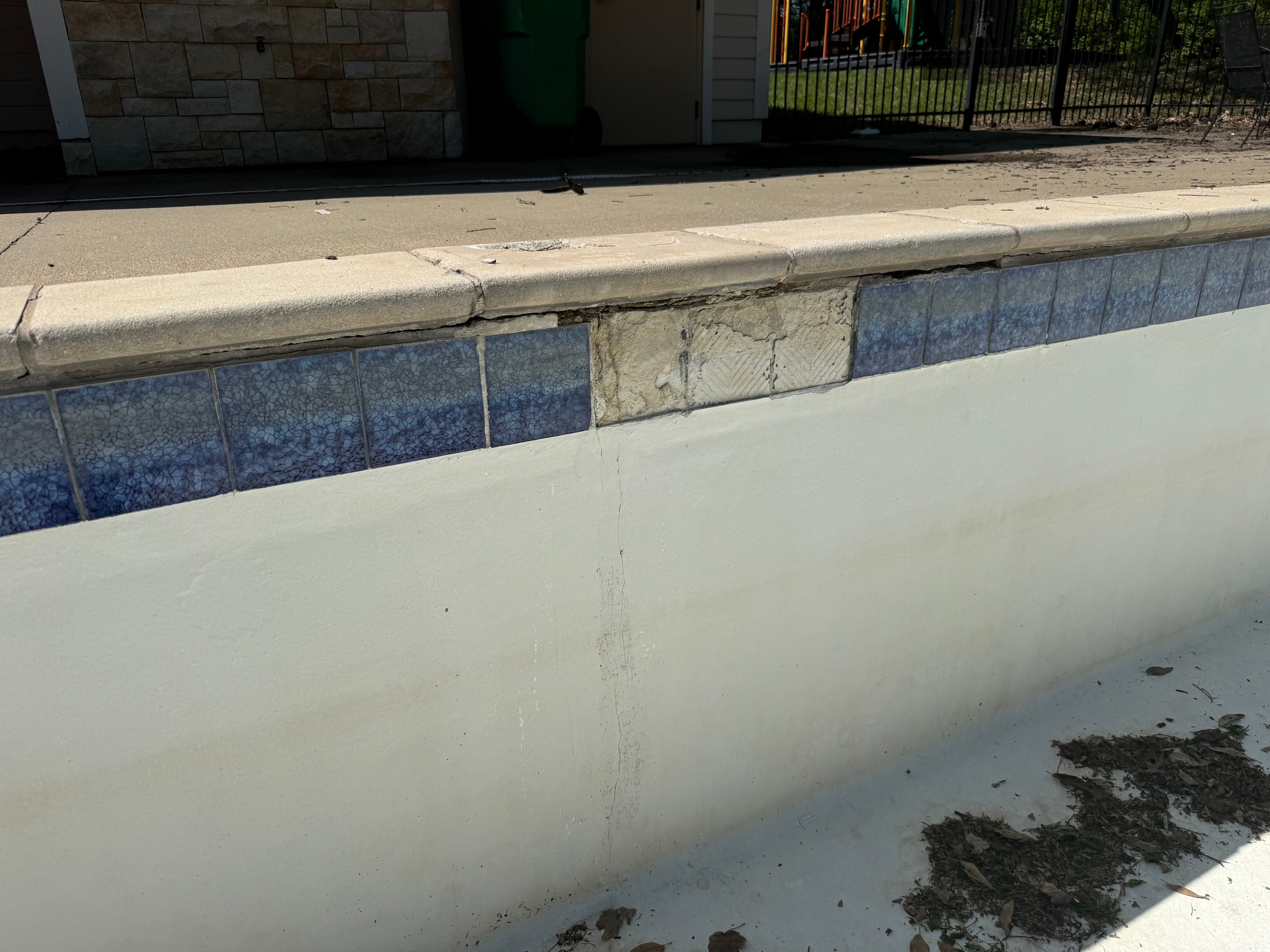 An empty, white plaster swimming pool shows visible damage near the coping